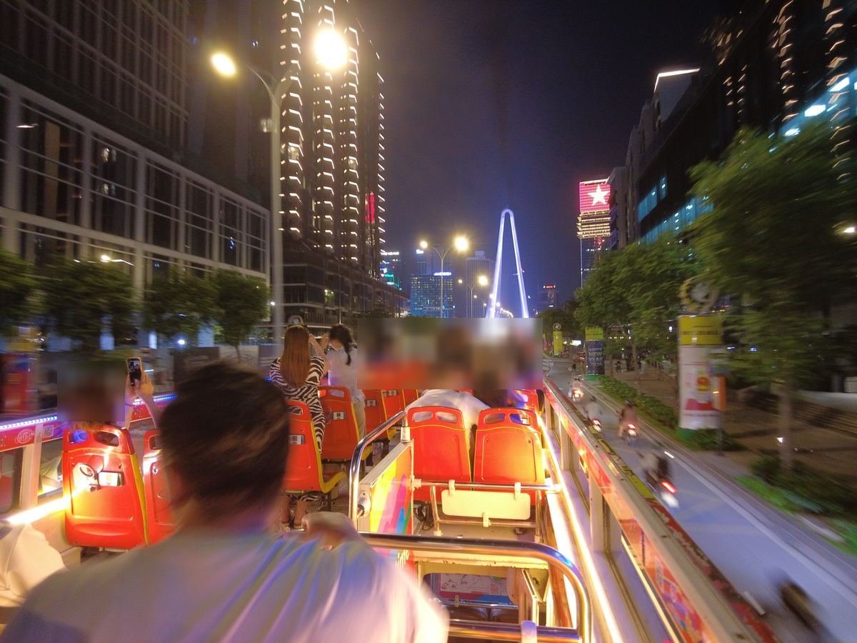 夜の大通りを走るルーフトップバスの2階席。正面にライトアップされたバーソン橋の主塔